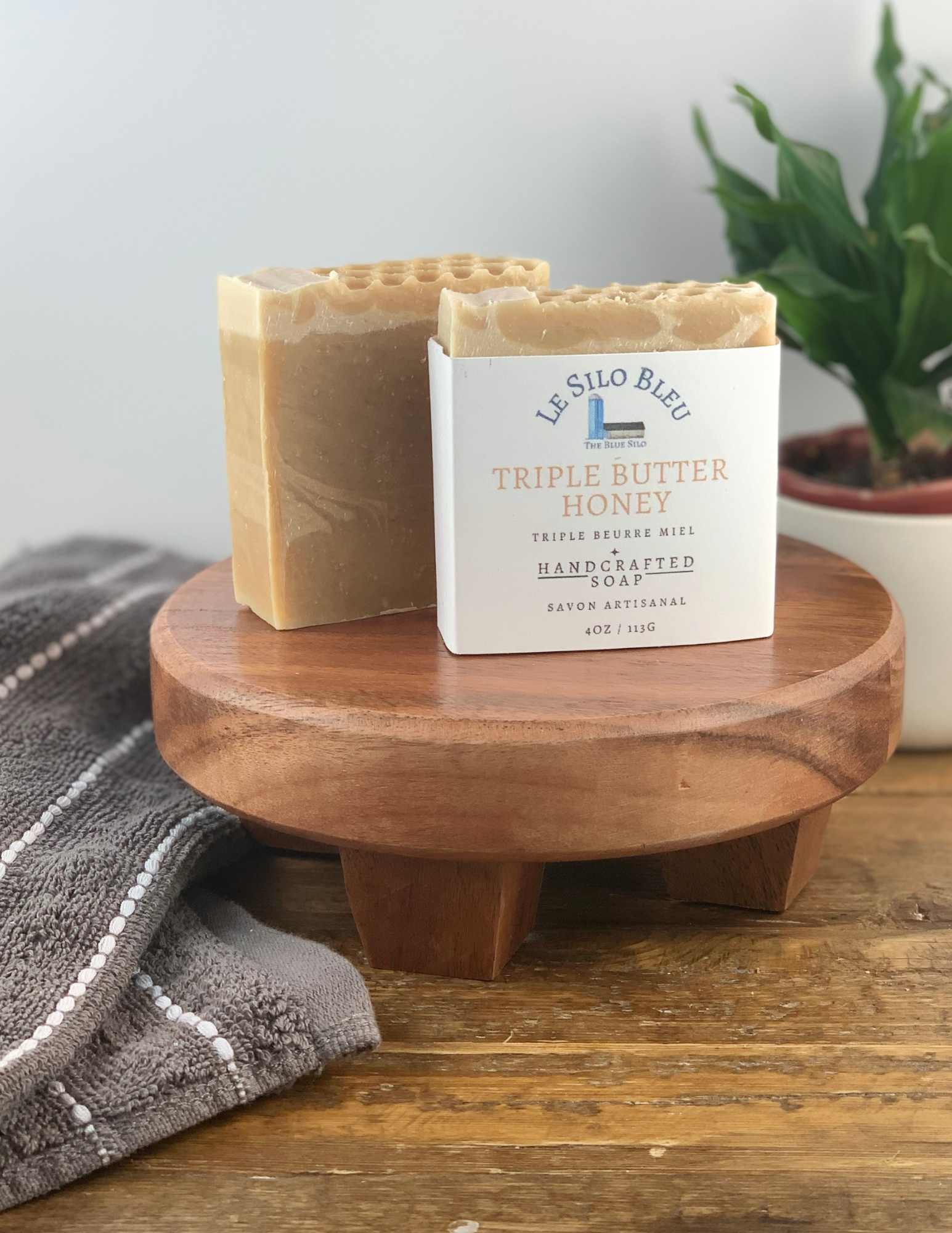 Two bars of Triple Butter Honey Soap sitting on a wooden platform with one soap wrapped in white labeling and one without. There is a green plant in a white pot in the background and a grey and white striped towel in the foreground. There is a wooden table and white background.