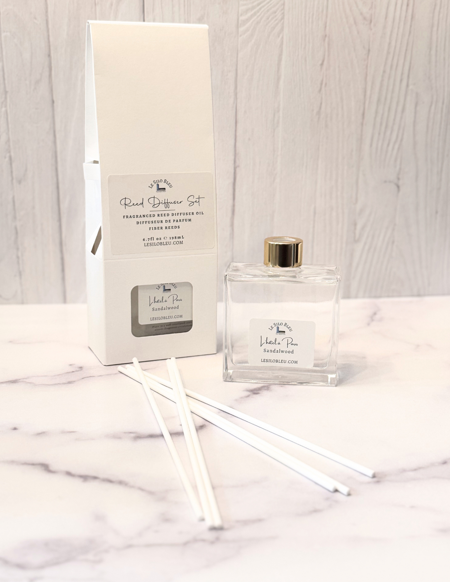 Reed diffuser set showing the bottle and reeds included in the package. All sitting on a white marble table with a white wood background.