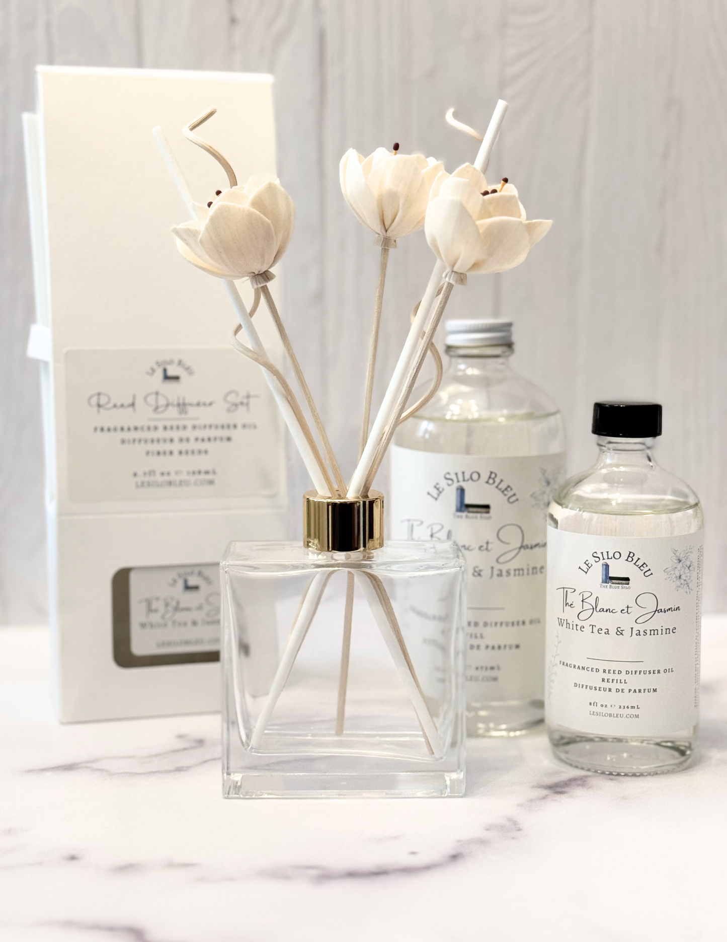 White Tea Scented Reed Diffuser set and collection showcasing the optional floral reeds along with the included white reeds and additional for purchase bottles of 8oz or 16oz fragrance refills. The whole scent collection is sitting on a white marble table with a white wood background.