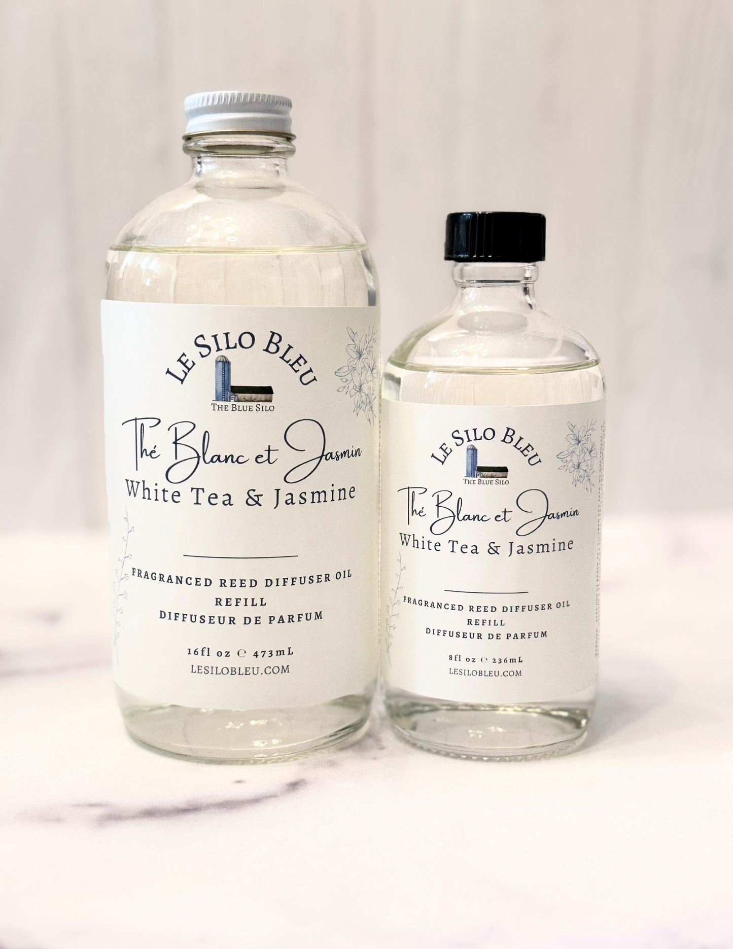 White Tea and Jasmine Reed Diffuser Refill bottles in 8oz and 16oz glass bottles are shown sitting on a white marble table with a white wood background. The cap for the large bottle is white and the cap for the small bottle is black.