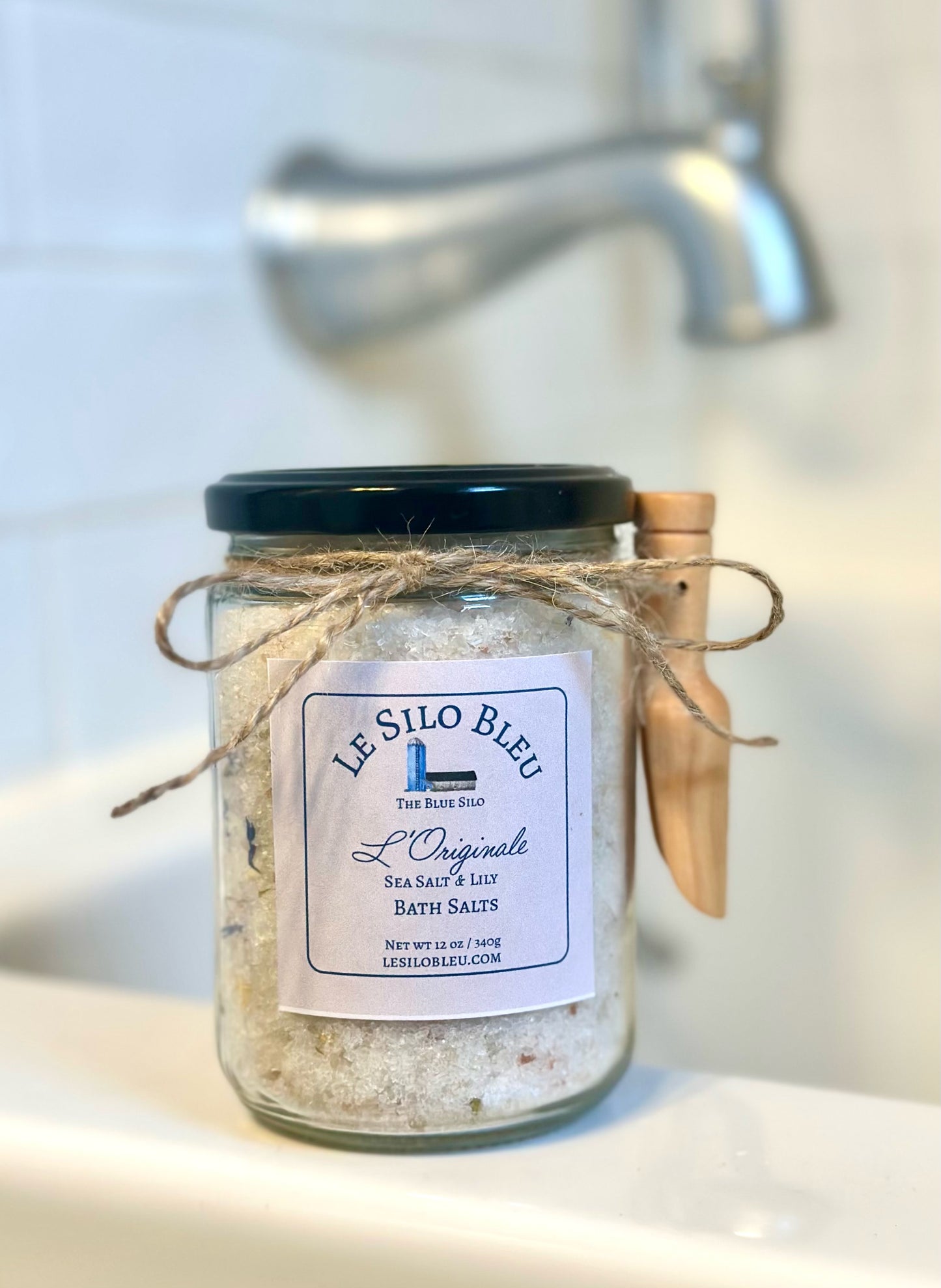 A jar of Sea Salt and Lily Bath salts sits on the edge of the bathtub. Details shows the jar tied with twine and a small wooden scoop is attached. Tub Faucet is blurred in the backgound. 