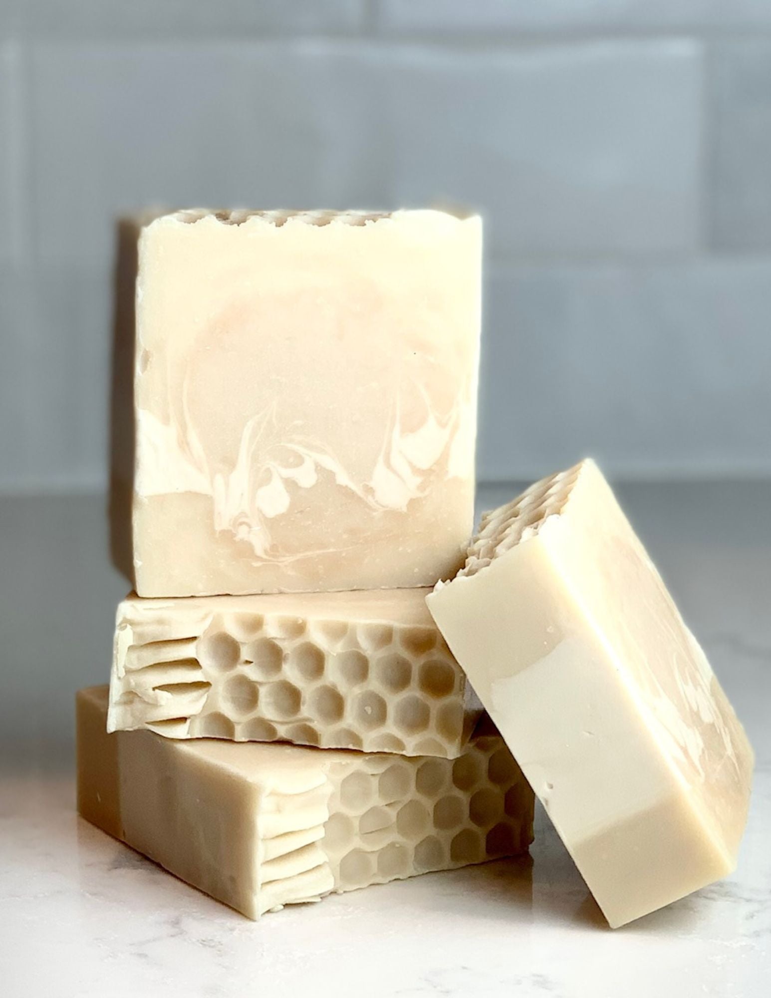 Triple Butter Honey soap stack of three with one leaning on stack detailing the honeycomb design on top and the white swirls in the honey colored soap. All are on a marble counter with a white tile backspash. 