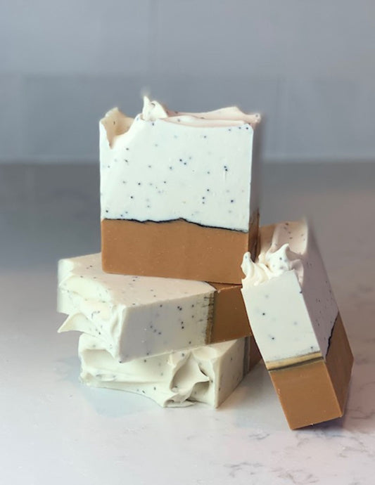 This fun bar of soap features a deep yellow orange bottom layer with a thin line of activated charcoal and a white creamy top with poppy seeds throughout.  Two bars are stacked with a third bar on top showing the details and a fourth bar is leaning against the stack. They sit on a marble counter with a white background. 