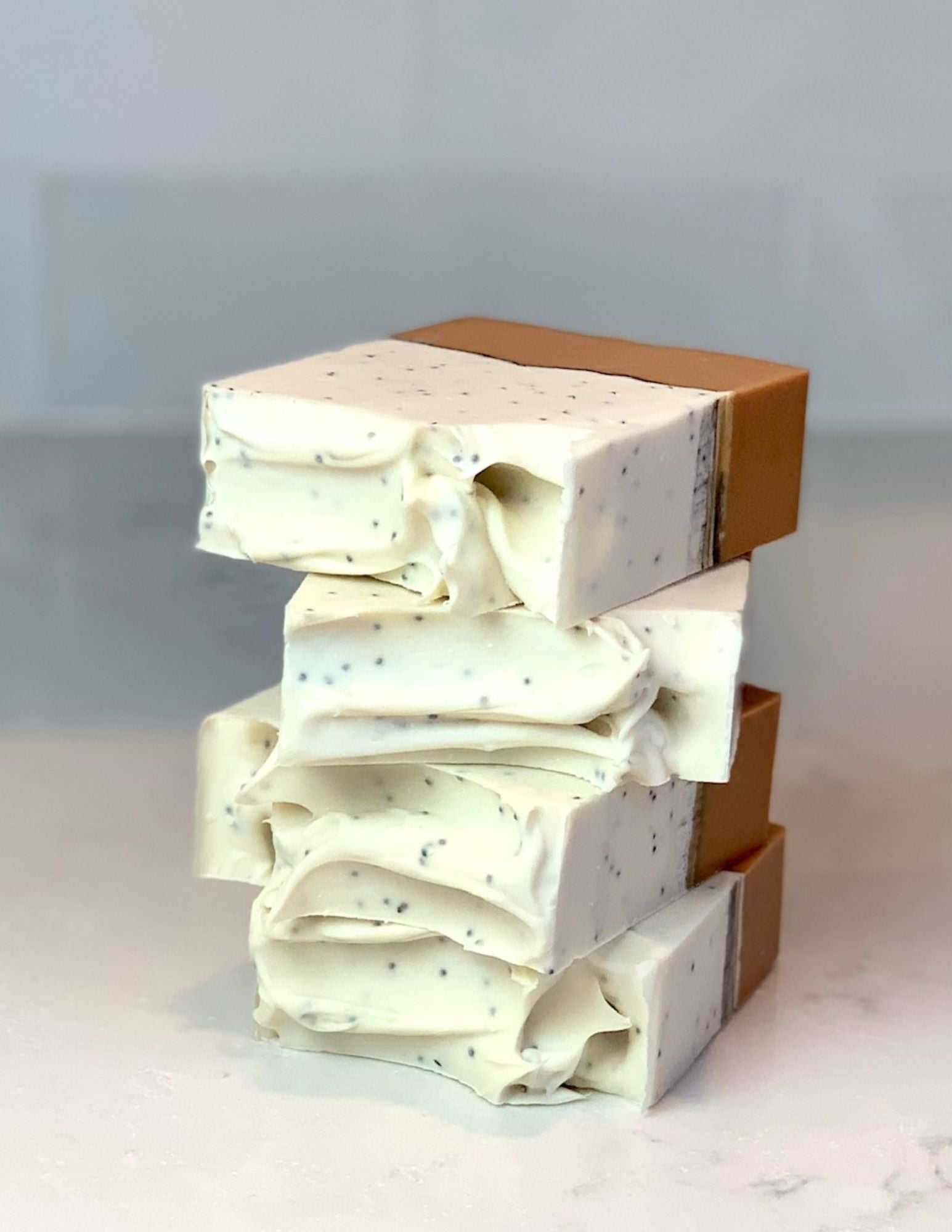 This fun bar of soap features a deep yellow orange bottom layer with a thin line of activated charcoal and a white creamy top with poppy seeds throughout.  Four bars are stacked. They sit on a marble counter with a white background. 