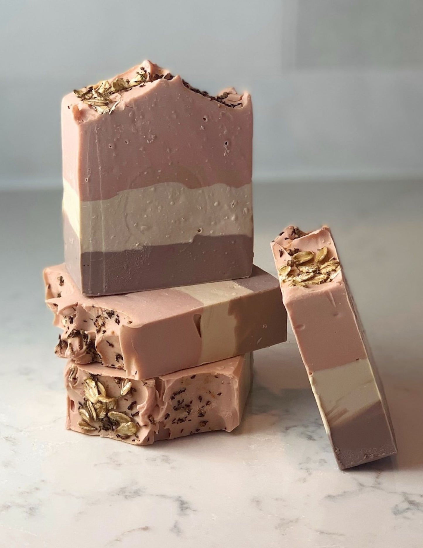 This handcrafted cold-processed soap is made with oat milk and natural clays. This lovely, oat-scented bar is extra creamy and produces a nice lather. A stack of three of these three color bars sits on a marble counter with another bar leaning against them. Made with Brazilian Purple Clay and Rose Clay for a pretty striped effect. Topped with Oats and Heather flowers.