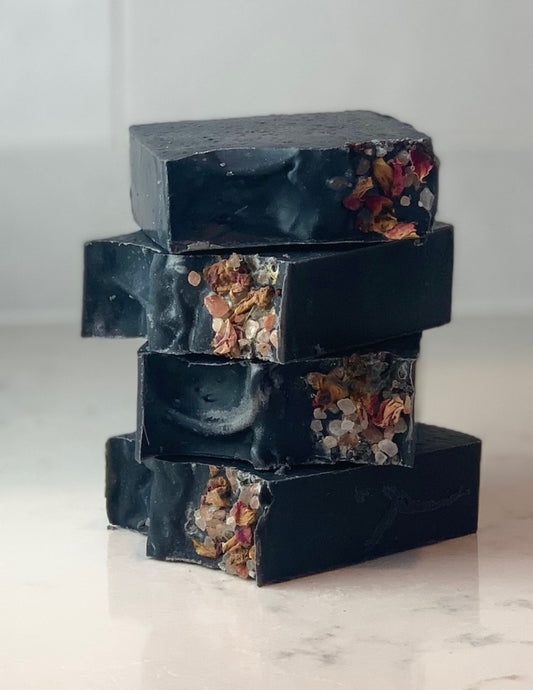This beautiful deep black soap with pink and red rose petals and pink sea salt are stacked four high on a marble table with a white background.
