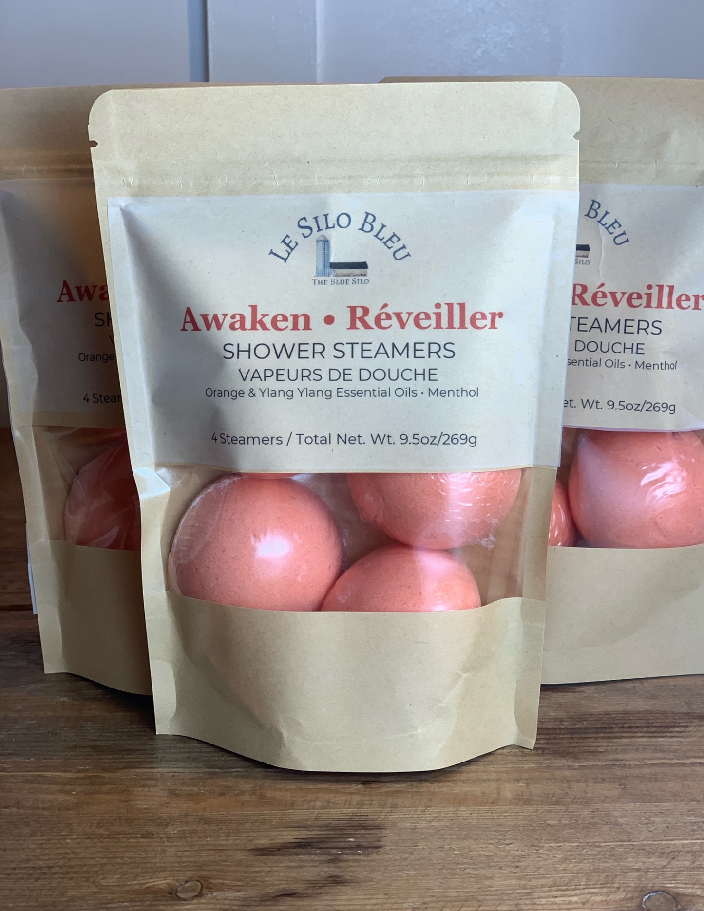 Three packages of Awaken Reveiller Shower Steamers sit on a wooden table with a white bead board background. Three orange steamers are visible with the package windows.