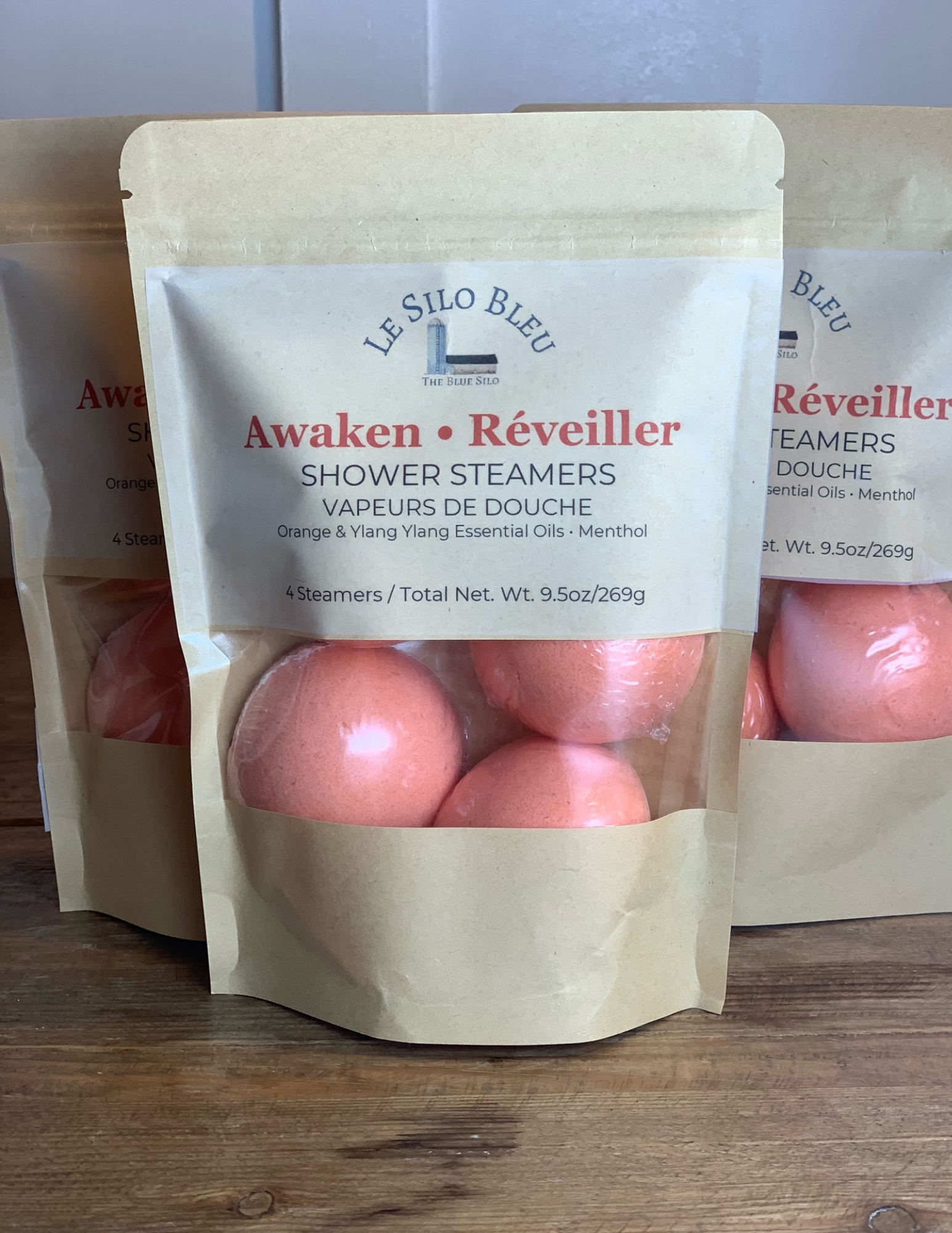Three packages of Awaken Reveiller Shower Steamers sit on a wooden table with a white bead board background. Three orange steamers are visible with the package windows.