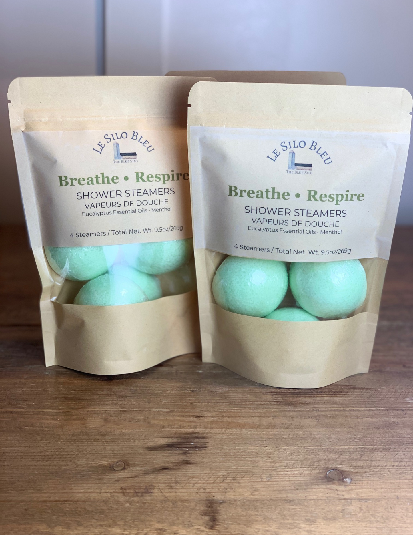 Two packages of Breathe, Respire Shower Steamers sit on a wooden table with a white bead board background. Three green steamers are visible with the package windows.
