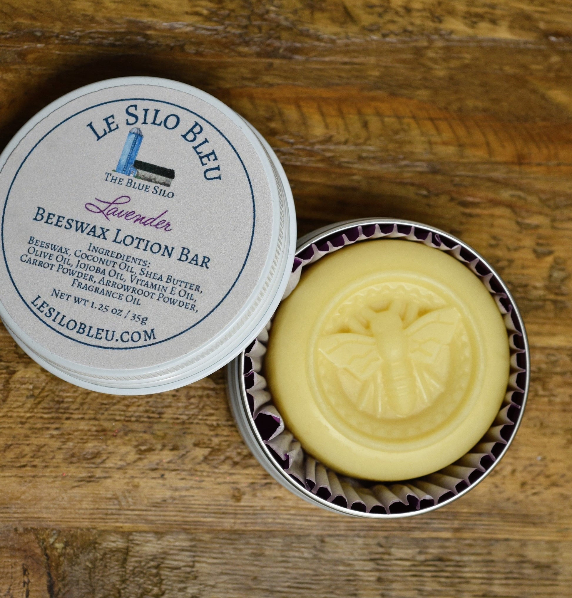 Lavender Beeswax Lotion Bar Tin 1.25oz showing bar with bee shape emblem. The open white tin is showing the yellowish round bar of lotion and they are laying on a wooden table.