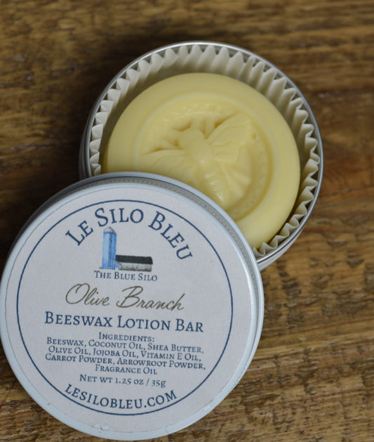 Olive Scented Beeswax Lotion Bar Tin 1.25oz showing bee shape emblem on round yellowish bar. This round tin is open and laying on a wooden table.
