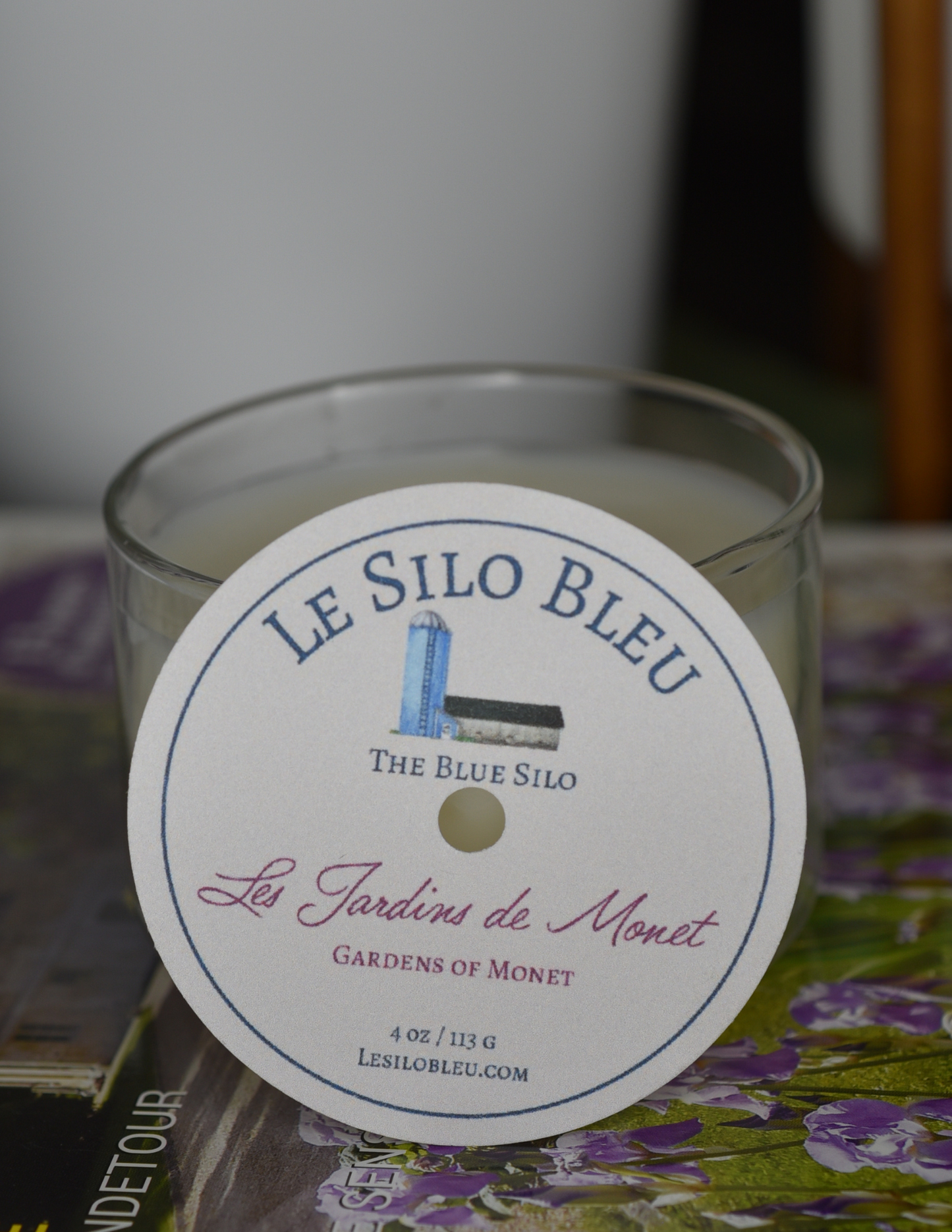 Les Jardins de Monet, a floral  scented candle is sitting on top of a French magazine with a plant in a white pot in the background.