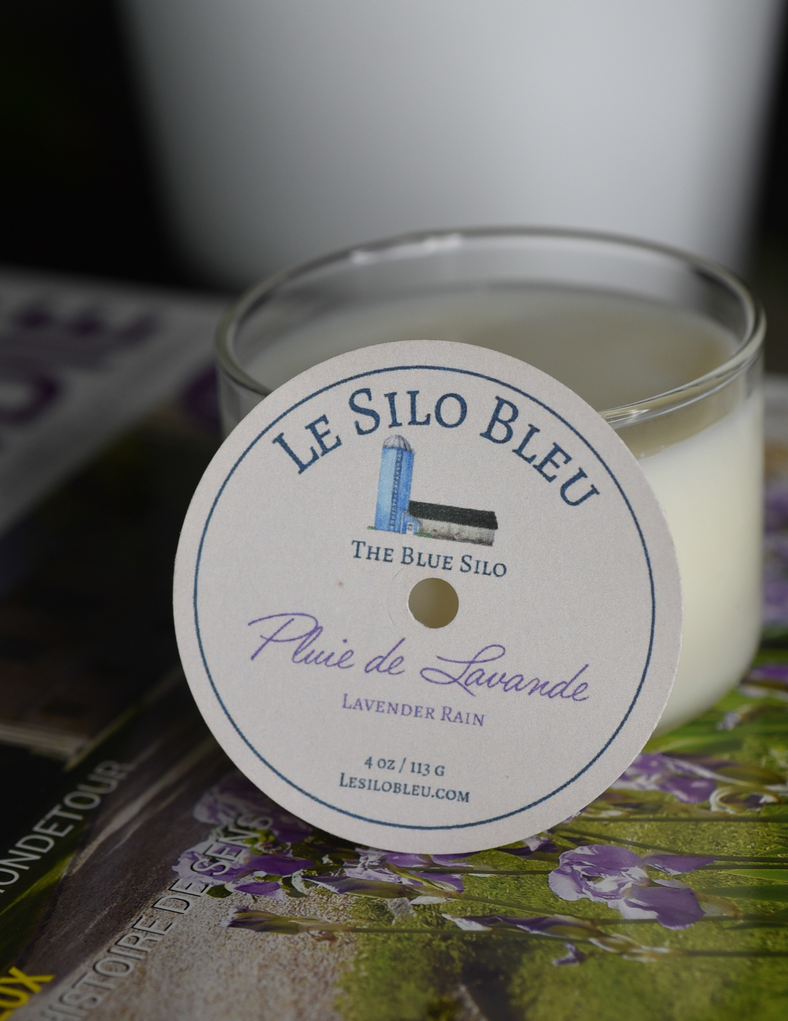A lavender and rain scented candle is sitting on top of a French magazine with a plant in a white pot in the background.