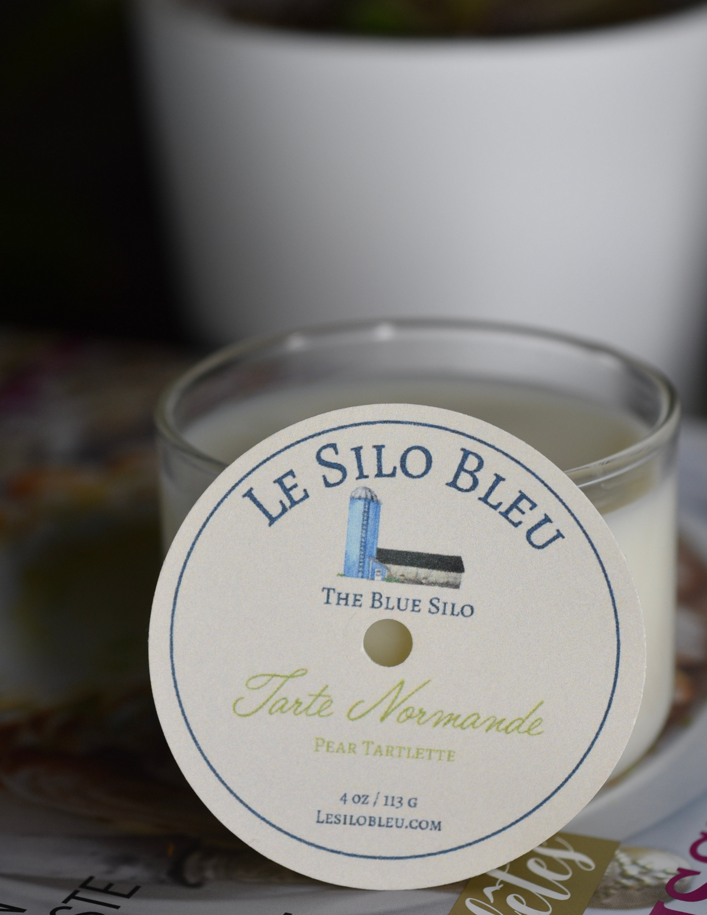 A pear and almond scented candle is sitting on top of a French magazine with a plant in a white pot in the background.