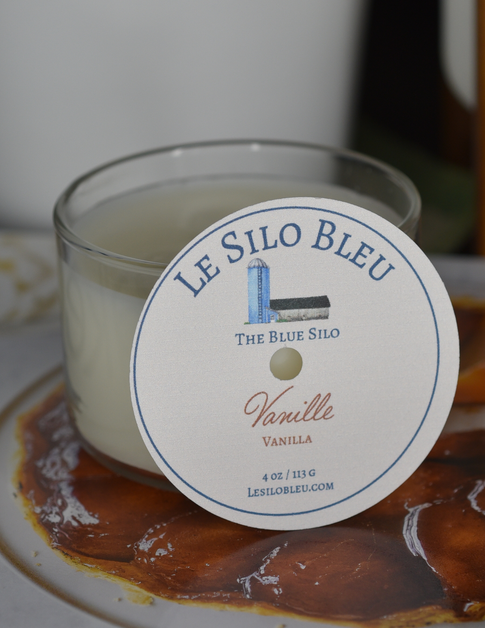 A vanilla  scented candle is sitting on top of a French magazine with a plant in a white pot in the background.