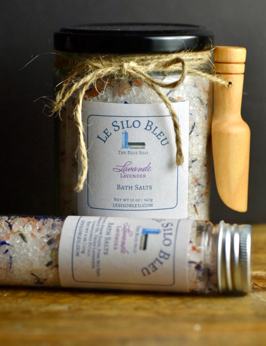 Lavender Bath Salts 12 oz Jar with small wooden scoop tied to jar with twine and a 4 oz Tube with metal top. They are sitting on a wood table with a black background.