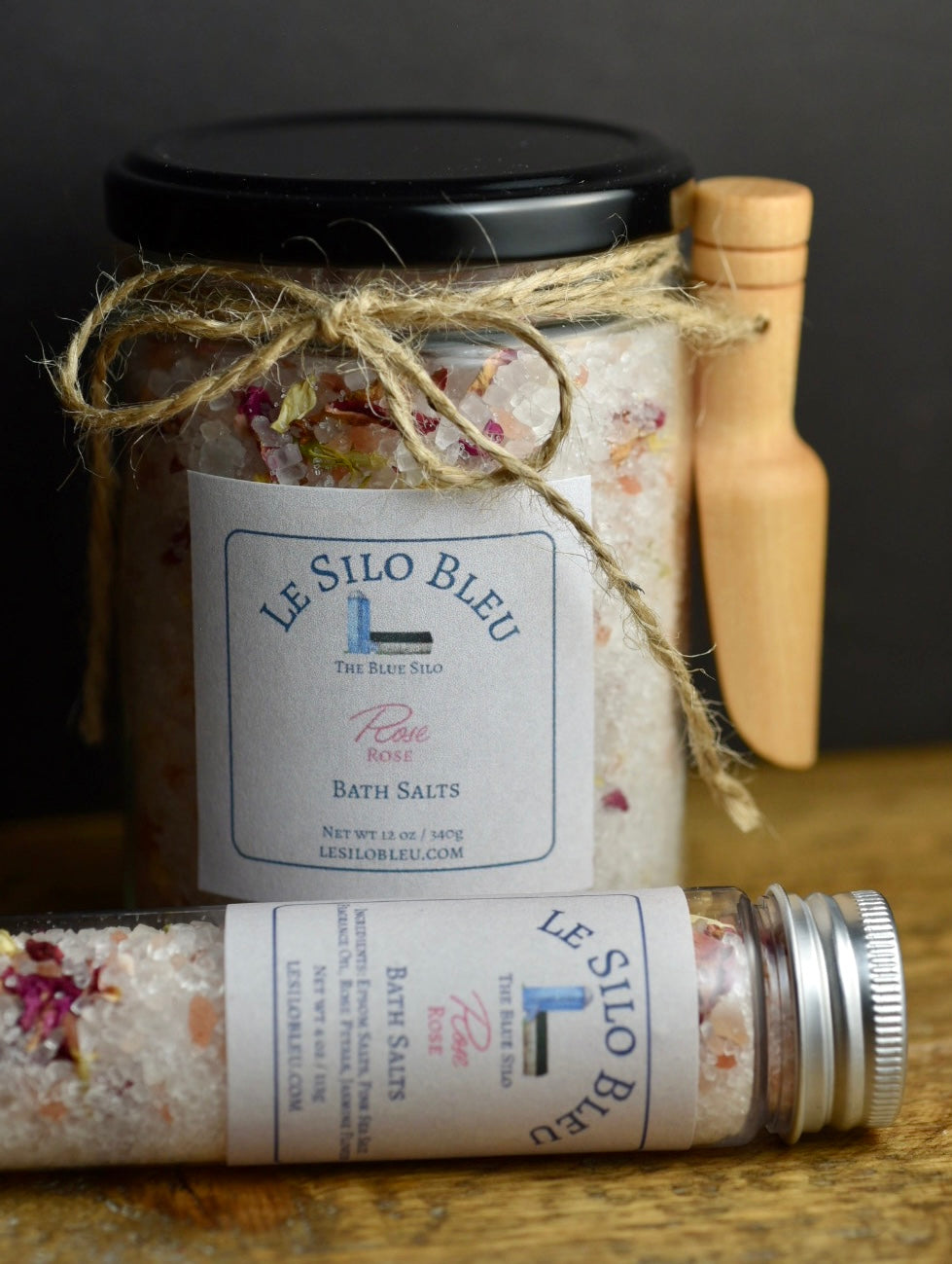Rose Bath Salts 12 oz Jar with small wooden scoop attached with twine and a bow and 4 oz Tube with metal top. They are laying on a wooden table with a black background.