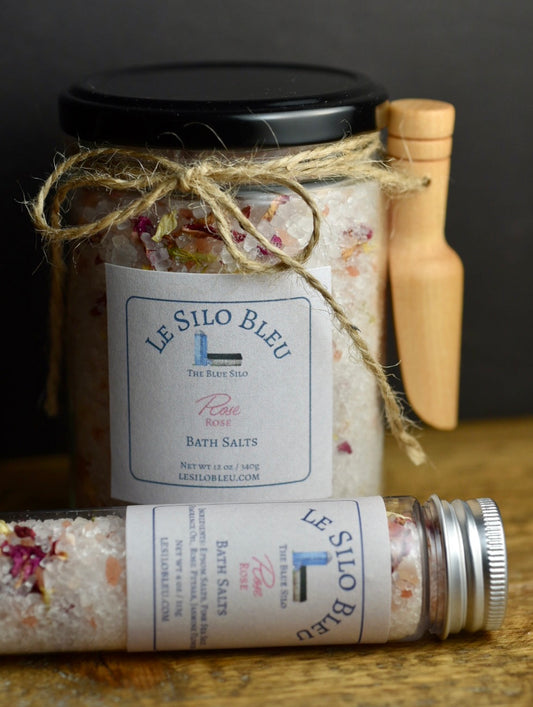 Rose  Bath Salts 12 oz Jar with small wooden scoop attached with twine and a bow and 4 oz Tube with metal top. They are laying on a wooden table with a black background.