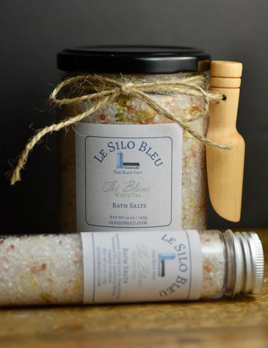White Tea Bath Salts 12 oz Jar with small wooden scoop attached by twine tied in a bow and 4 oz Tube with a metal lid sitting on a wooden table with a black background