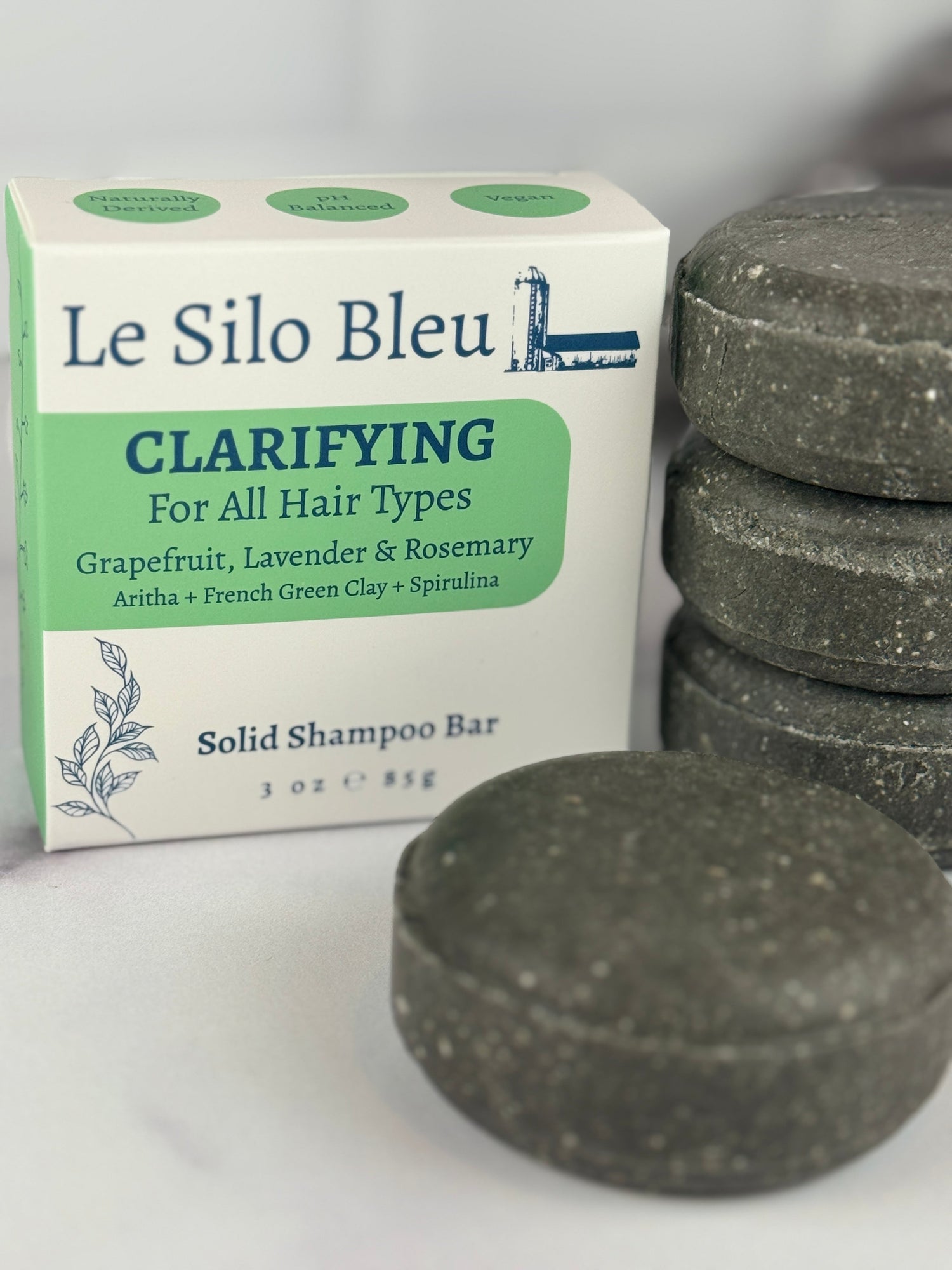  A stack of three with a single bar in front of the Clarifying solid shampoo package. Sitting on a white marble table with a white background. These bars are a rich deep dark green. 
