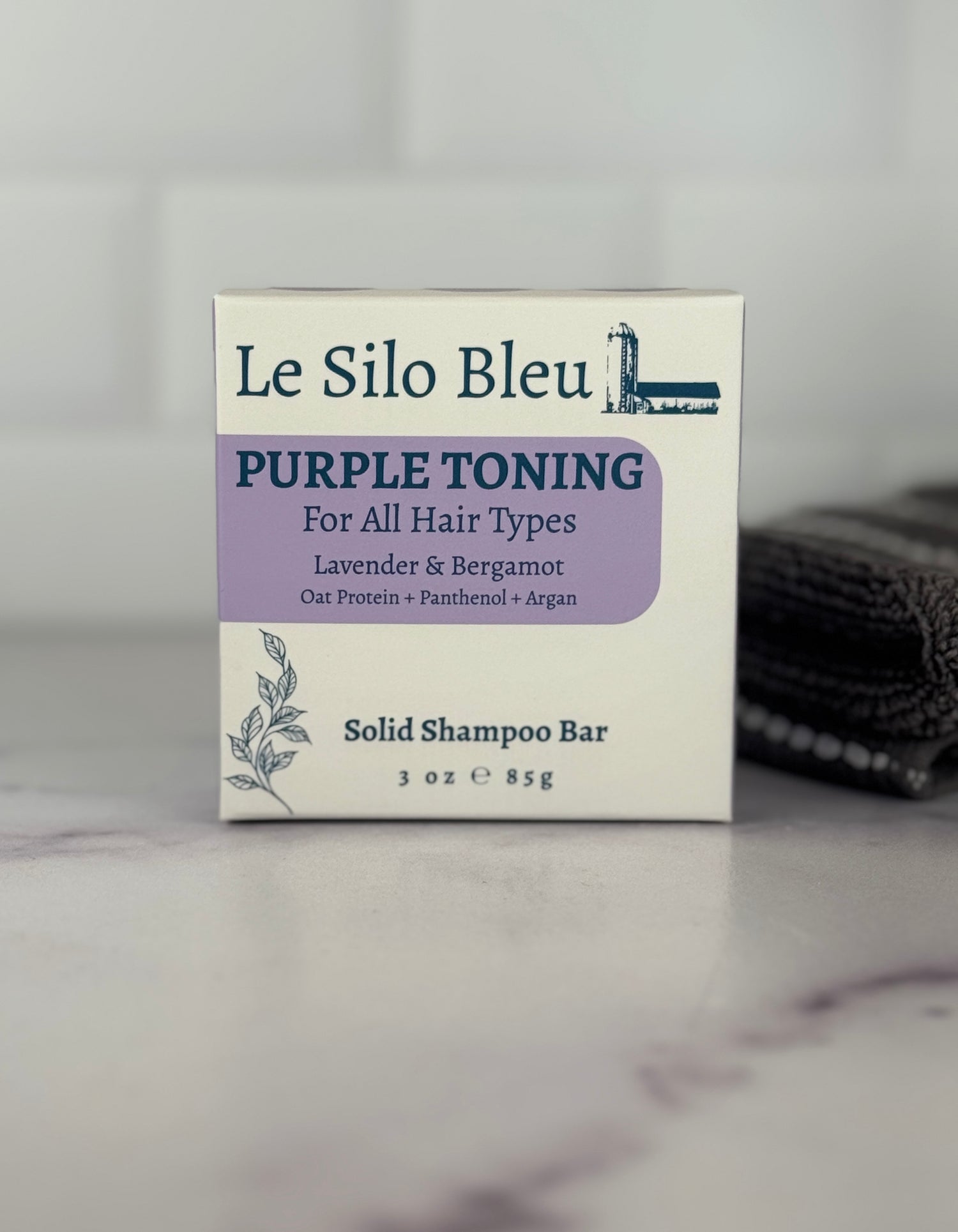Packaging for the purple toner solid shampoo bar sitting on a white and grey marble surface with a white backsplash and a grey and white stripped towel in the background.