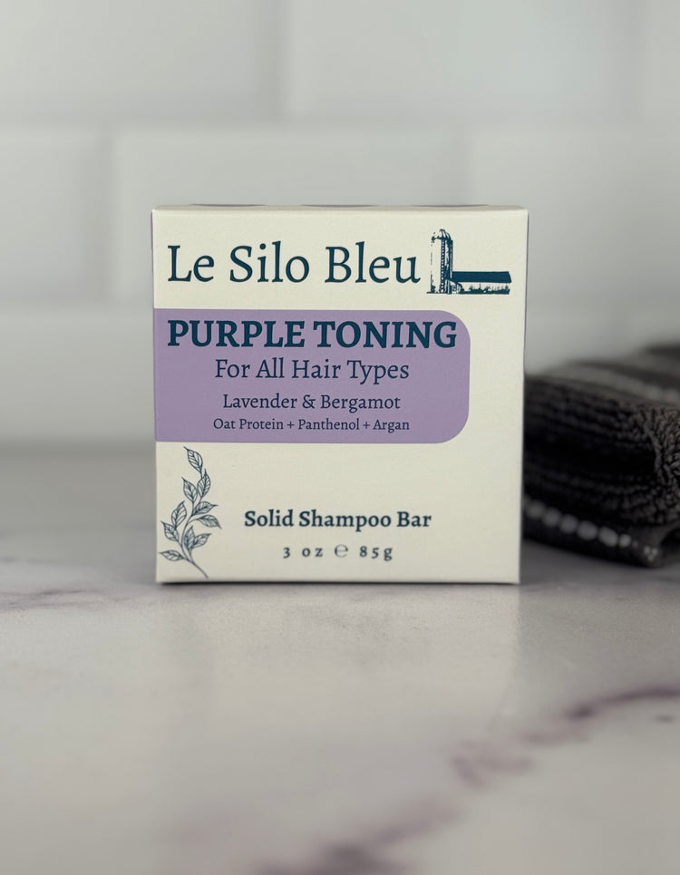 Packaging for the purple toner solid shampoo bar sitting on a white and grey marble surface with a white backsplash and a grey and white stripped towel in the background.