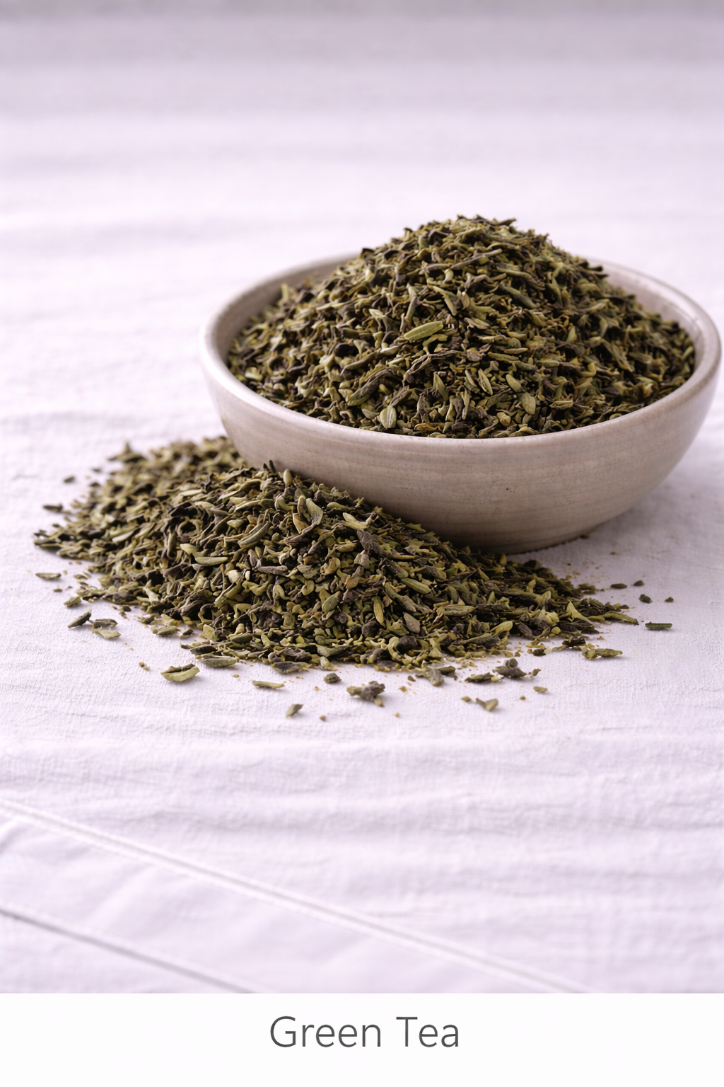 Dried green tea leaves in a rustic bowl with more leaves around it sitting on a linen tablecloth.