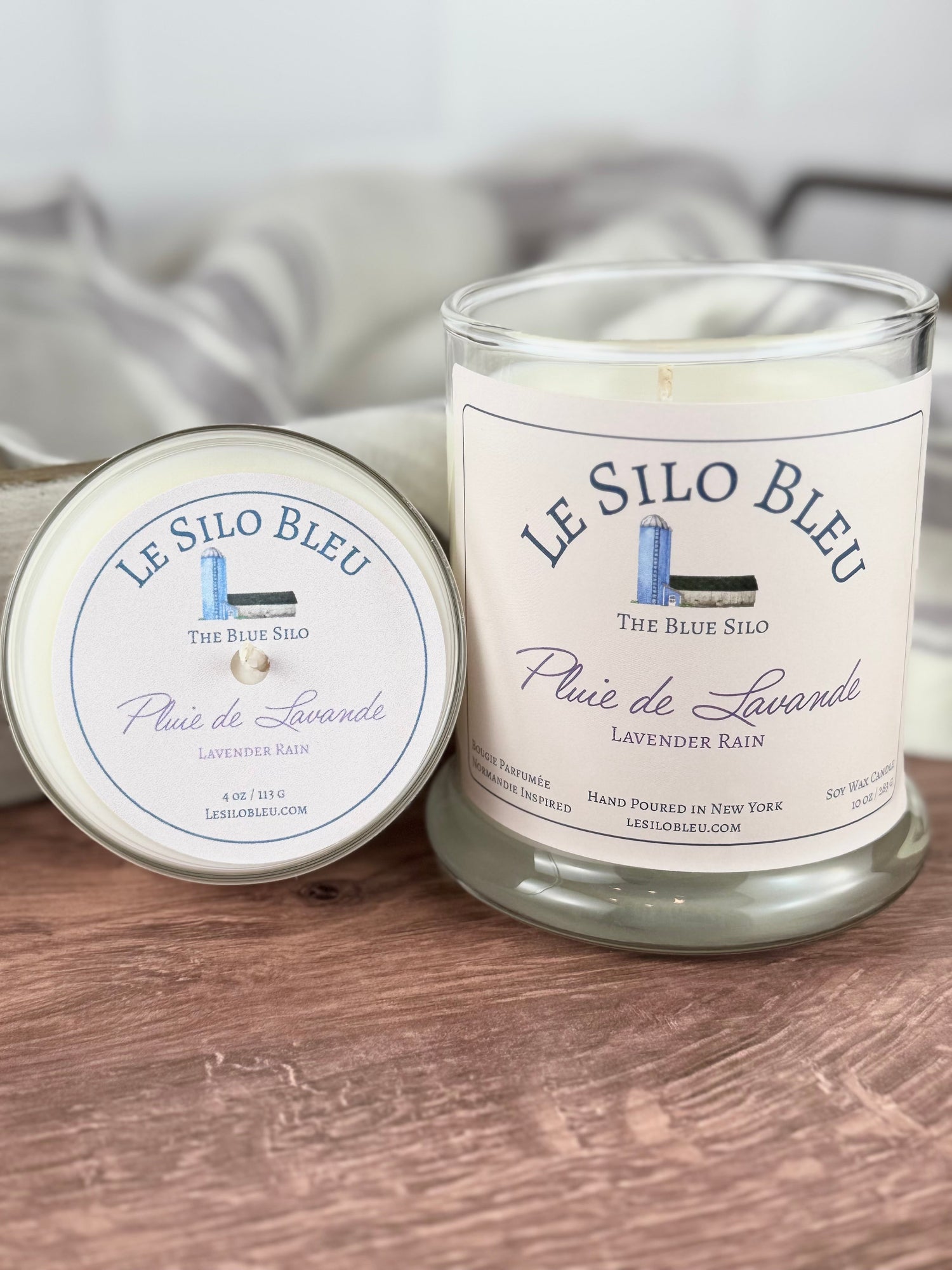 Two sizes of hand poured unlit soy wax candles in a glass jars labeled "Pluie de Lavande- Lavender Rain " sitting on a wooden tabletop. In the background there is a white linen towel with lavender stripes in an antique box with a metal handle.