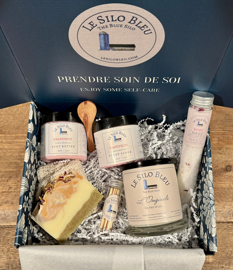 Le Silo Bleu self-care products with candle, bathsalts tube, handcrafted soap, and body butter sugar scrub set in a branded box sitting on a wooden surface.