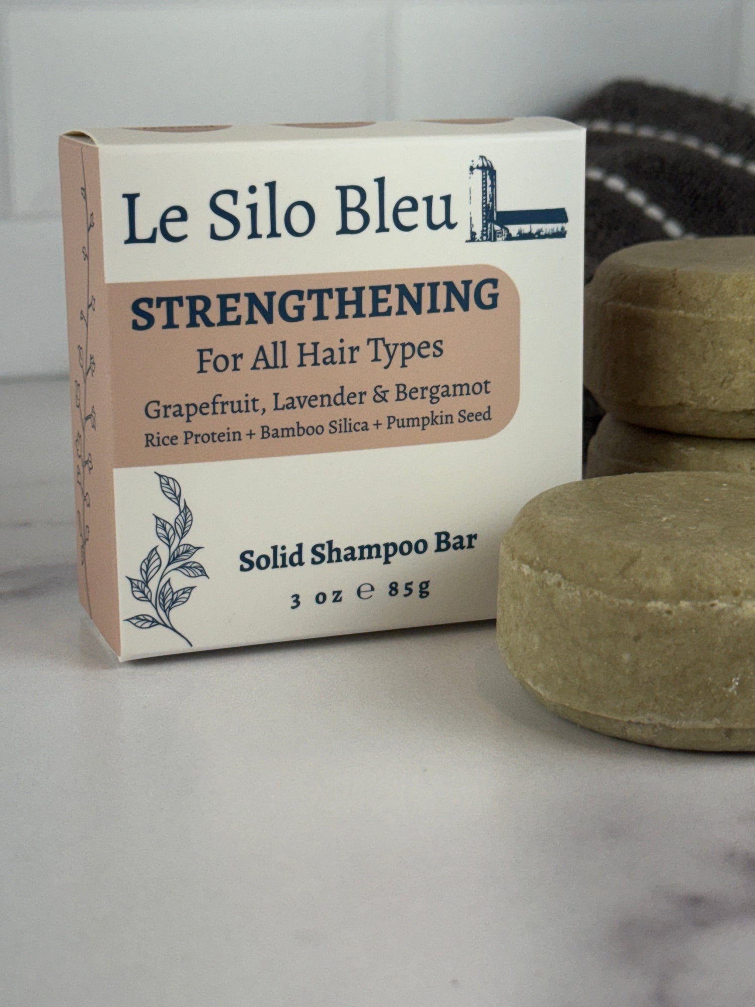 Single Strengthening solid shampoo bar with two stacked behind and the Le Silo Bleu packaging for the bars all on top of a white and grey marble surface with a white tiled backsplash. There is a folded grey and white striped towel in the background.