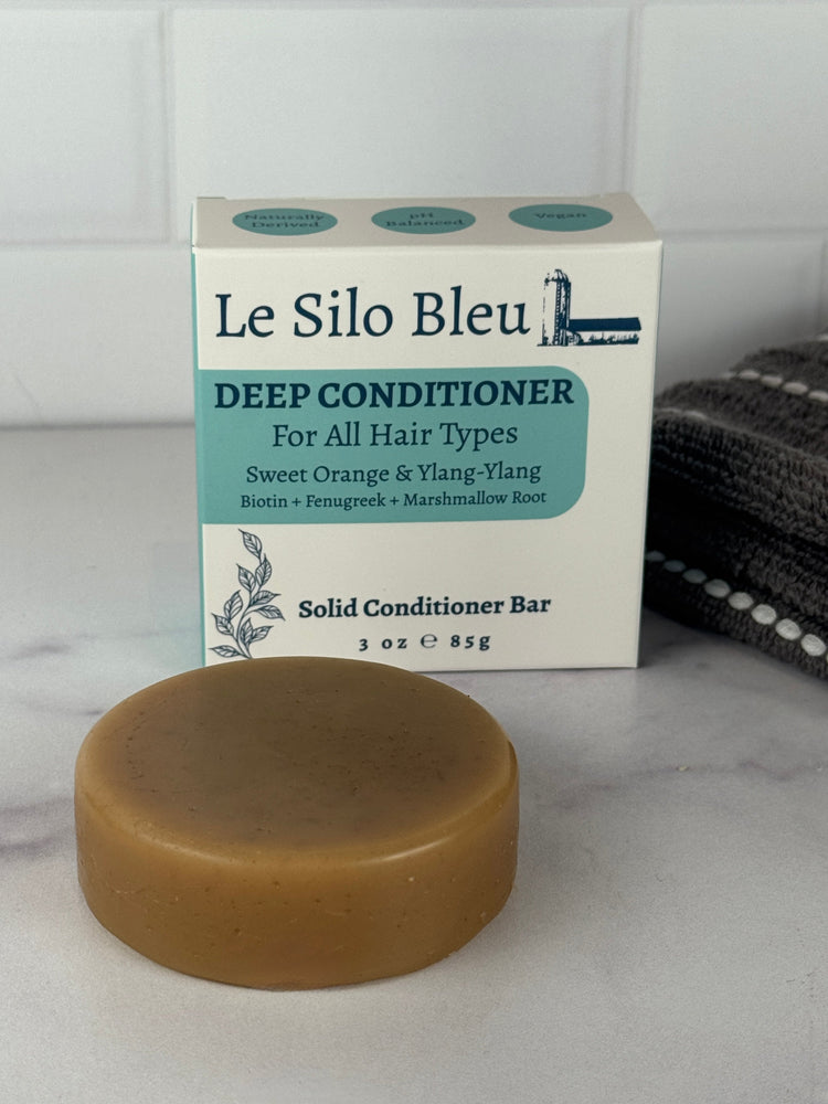 Single Deep Conditioner bar with the Le Silo Bleu packaging for the bars all on top of a white and grey marble surface with a white tiled backsplash. There is a folded grey and white striped towel in the background as well.
