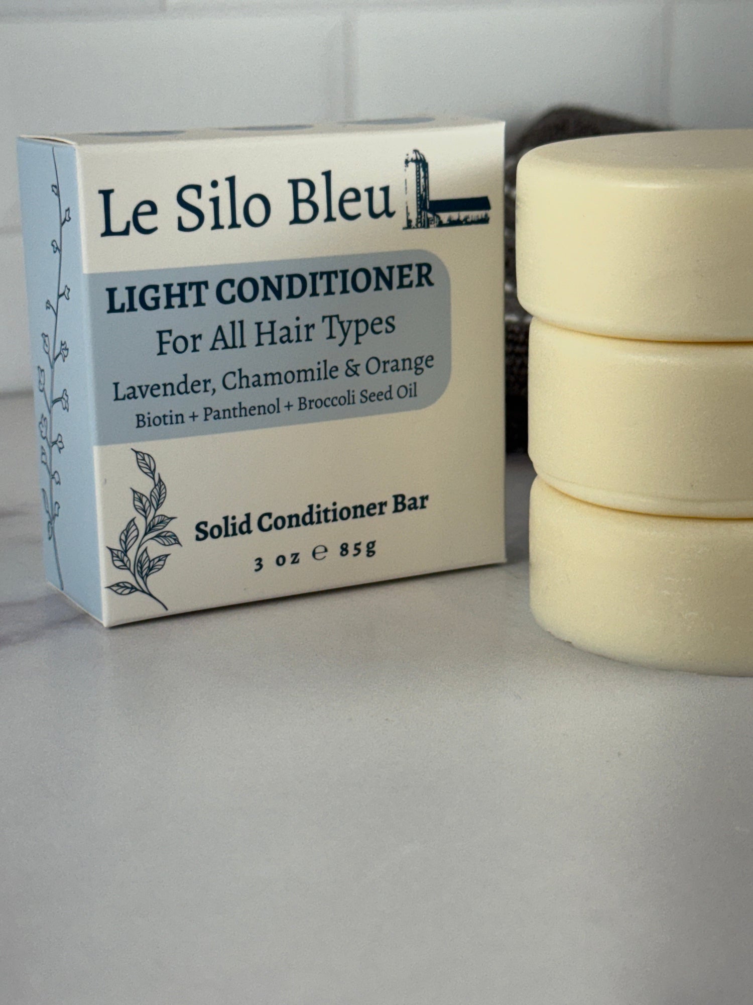 A stack of three cream colored Le Silo Bleu solid conditioner bars with their packaging sitting on a marble surface with a white tiled background.