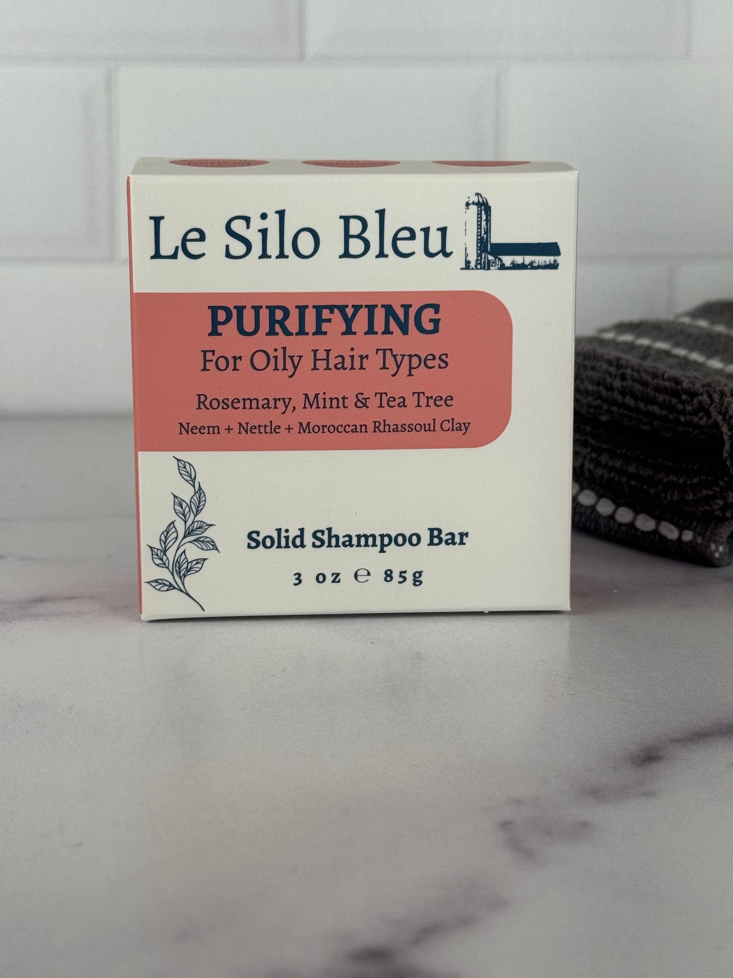 Purifying shampoo bar box sitting on a marble table with a white tile background and a grey towel on the table