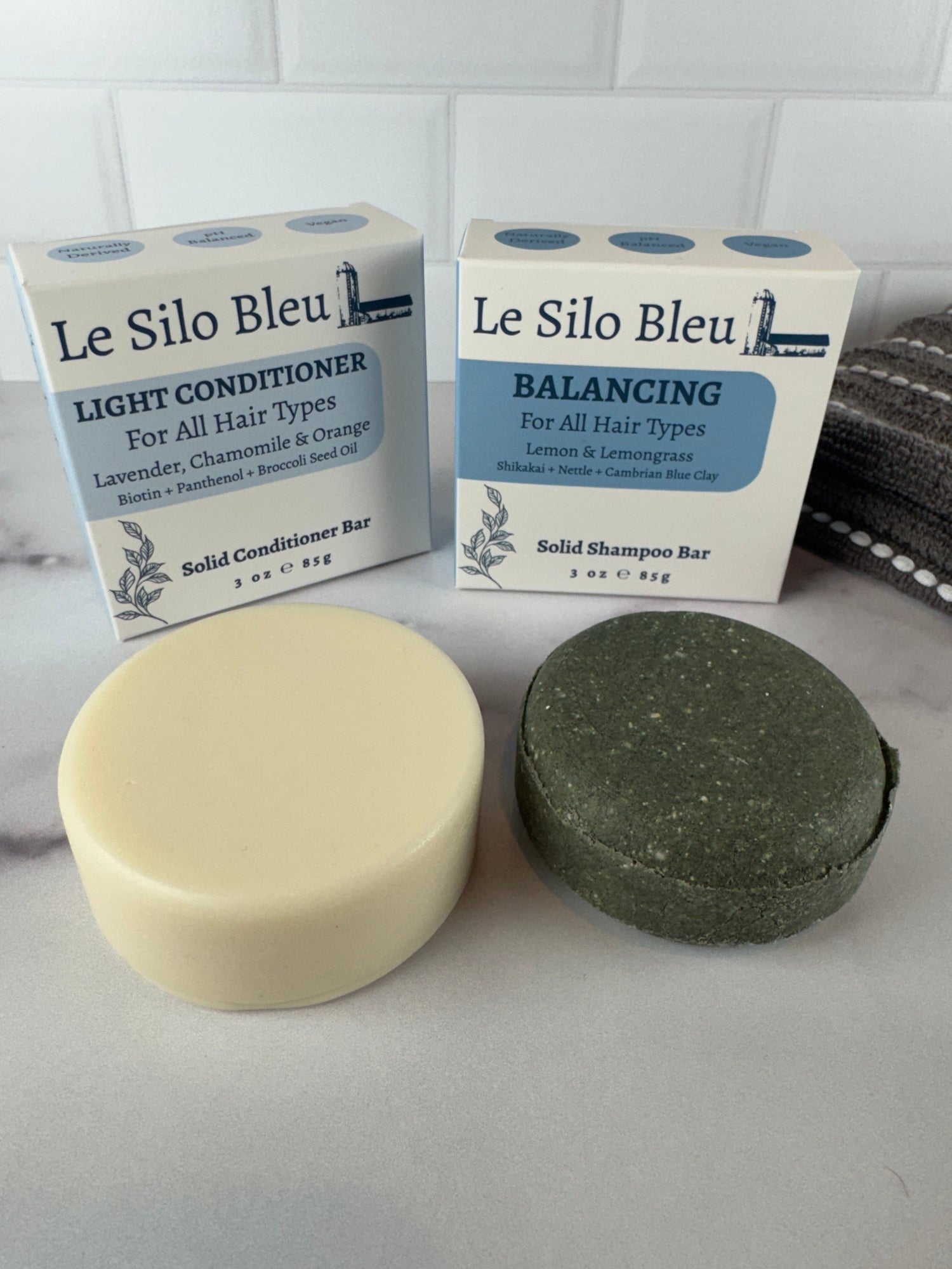 Balancing shampoo and Light conditioner bars with their packaging on a white table with a white tile background and grey towel sitting in the background.