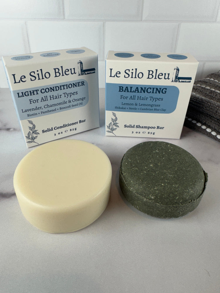 A light conditioner bar and a balancing solid shampoo bar sitting on a white marble table with a white tile backspash and a grey and white striped towel in the background. 