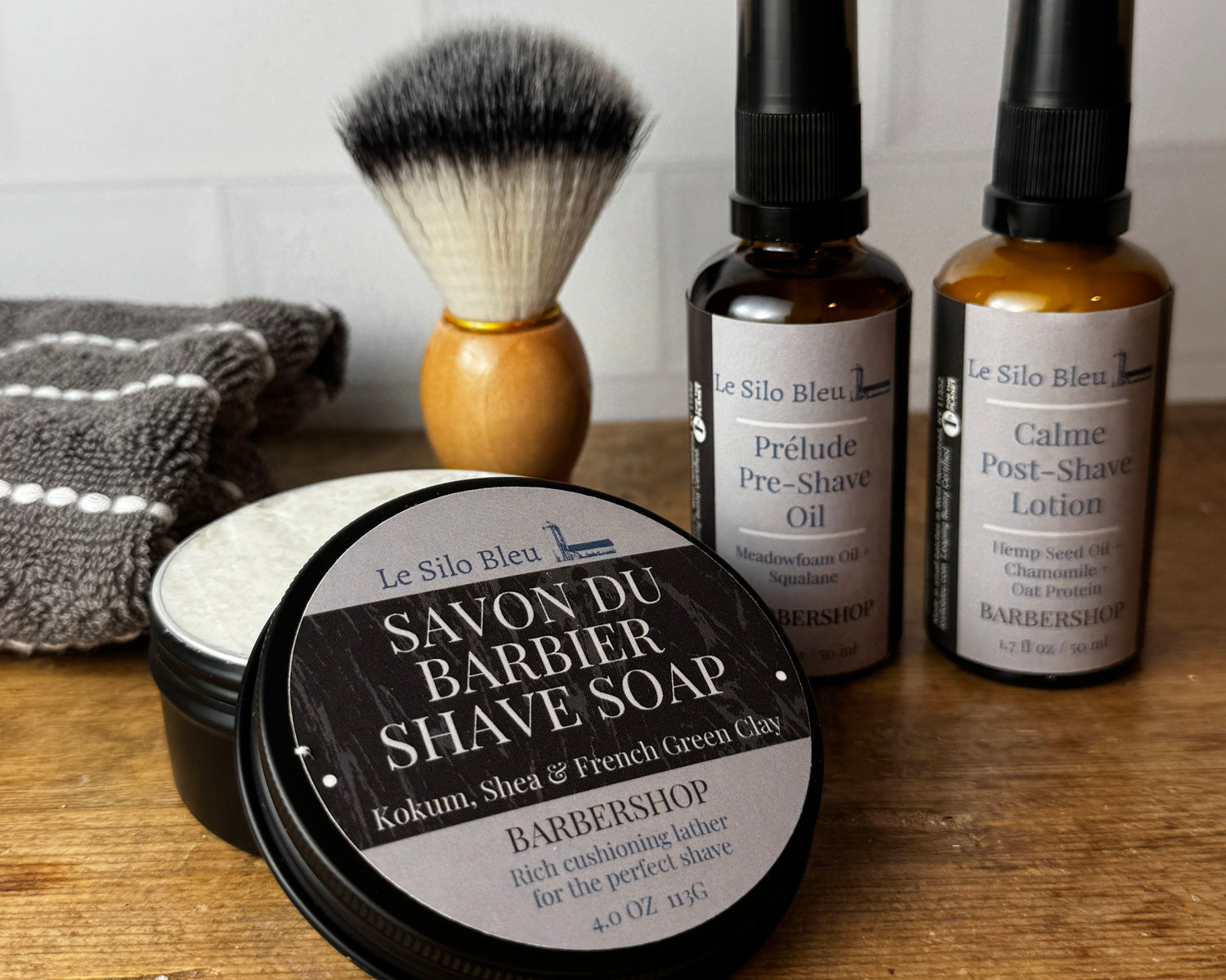 A men's grooming set arranged on a wooden surface against a white subway tile wall, including two amber bottles with black caps, a shaving brush with black and white bristles and a light wood handle, a black tin of Ultra Shave Soap, and a gray towel.