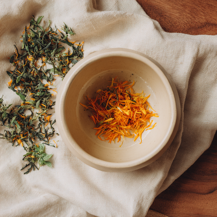 Calendula extract with flower materal in a ceramic bowl with plant matter on a linen tablecloth on a wooden table.