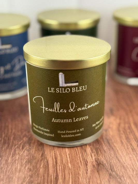 Candle jar labeled 'Le Silo Bleu Autumn Leaves' on a wooden surface with other jars in the background.