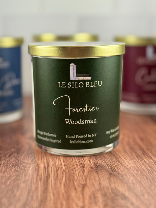 Le Silo Bleu candle labeled 'Forester Woodsman' on a wooden surface with other candles in the background.
