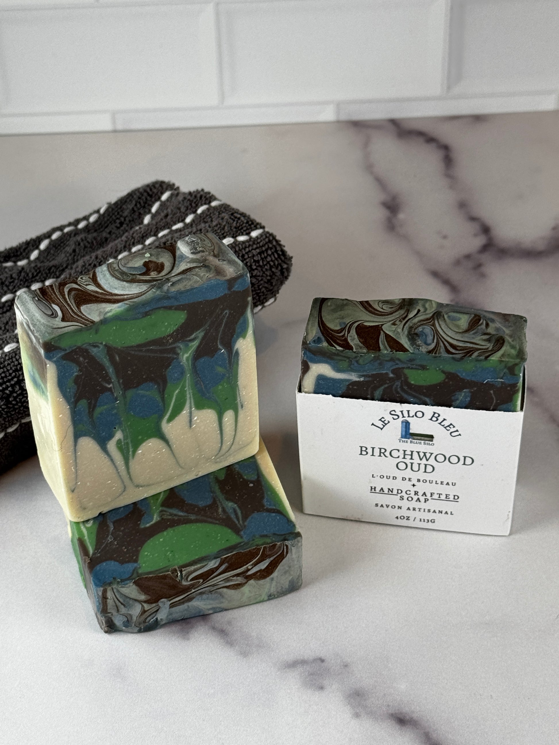 Two handmade soap bars with a marbled swirl pattern in green, blue, brown, and cream, displayed on a white marble surface next to a black-and-white striped cloth. One bar is standing upright with a white paper label that reads 'Silo Blue Birchwood'.