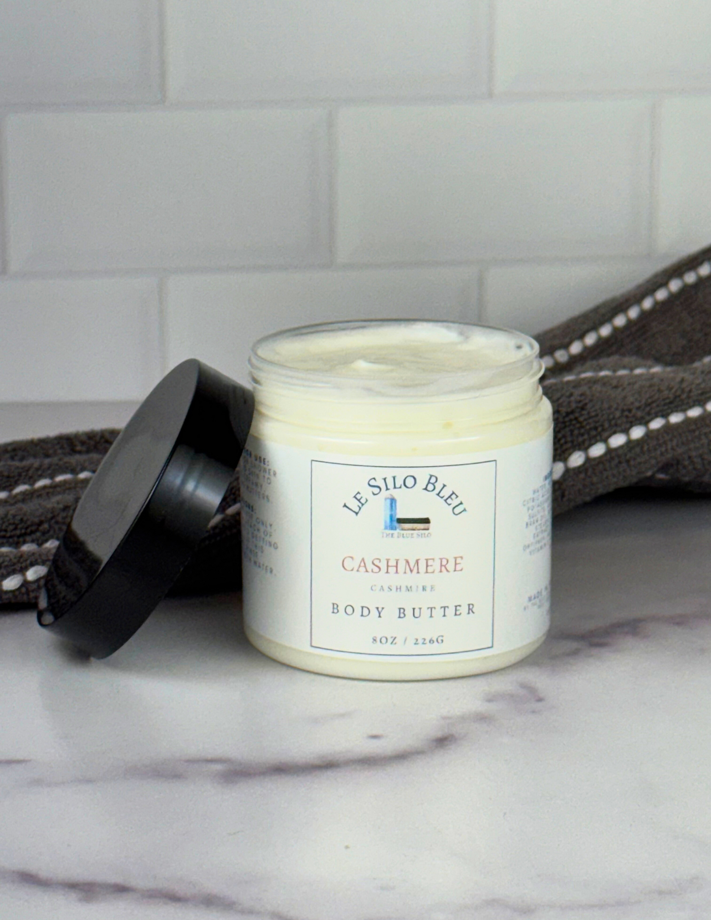 An open jar of Cashmere scented Body Butter with a black lid leaning against the jar sitting on top of a white marble table with a grey and white stripped towel in the background. The label shows the logo and the name of the scent. 