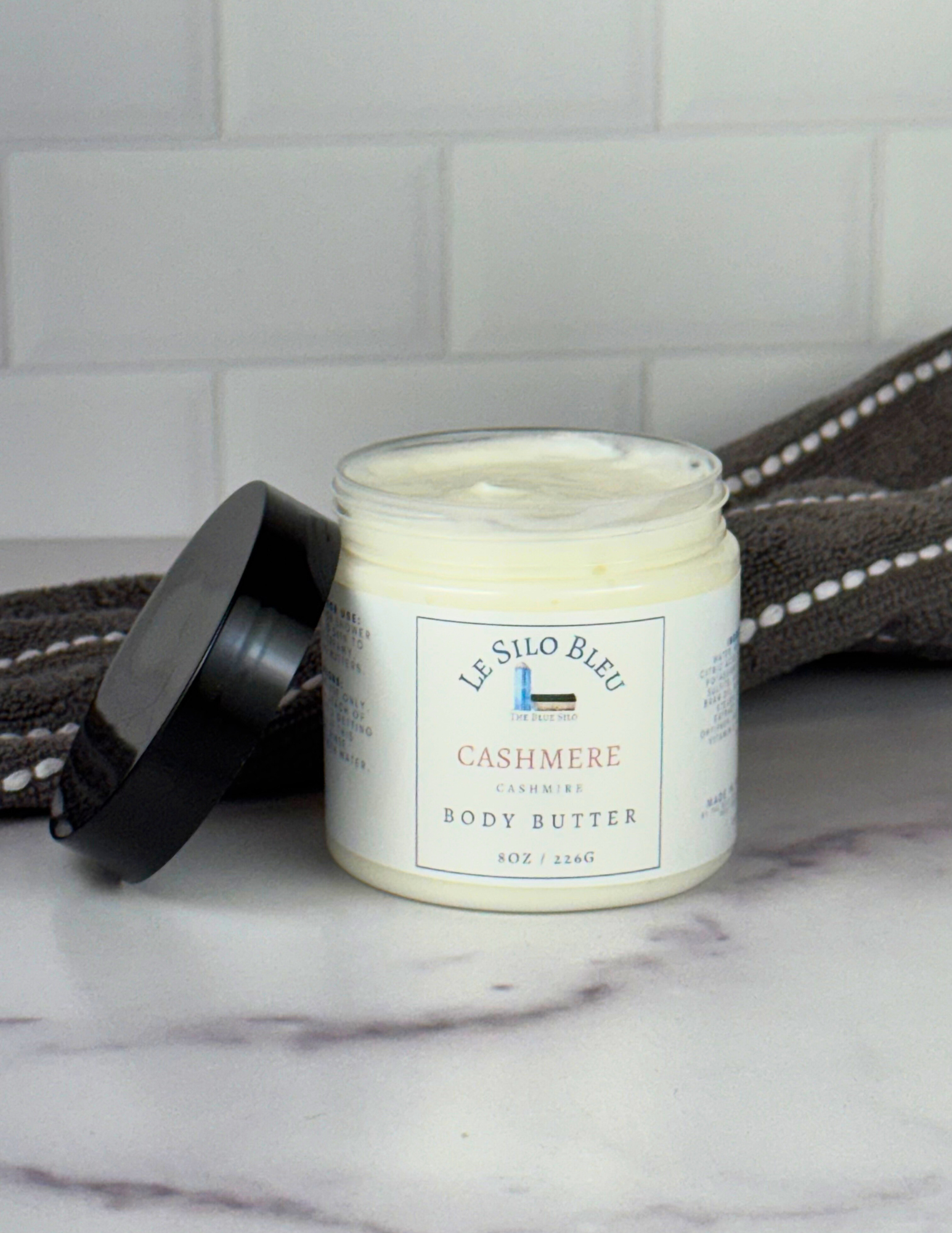 An open jar of Cashmere scented Body Butter with a black lid leaning against the jar sitting on top of a white marble table with a grey and white stripped towel in the background. The label shows the logo and the name of the scent. 