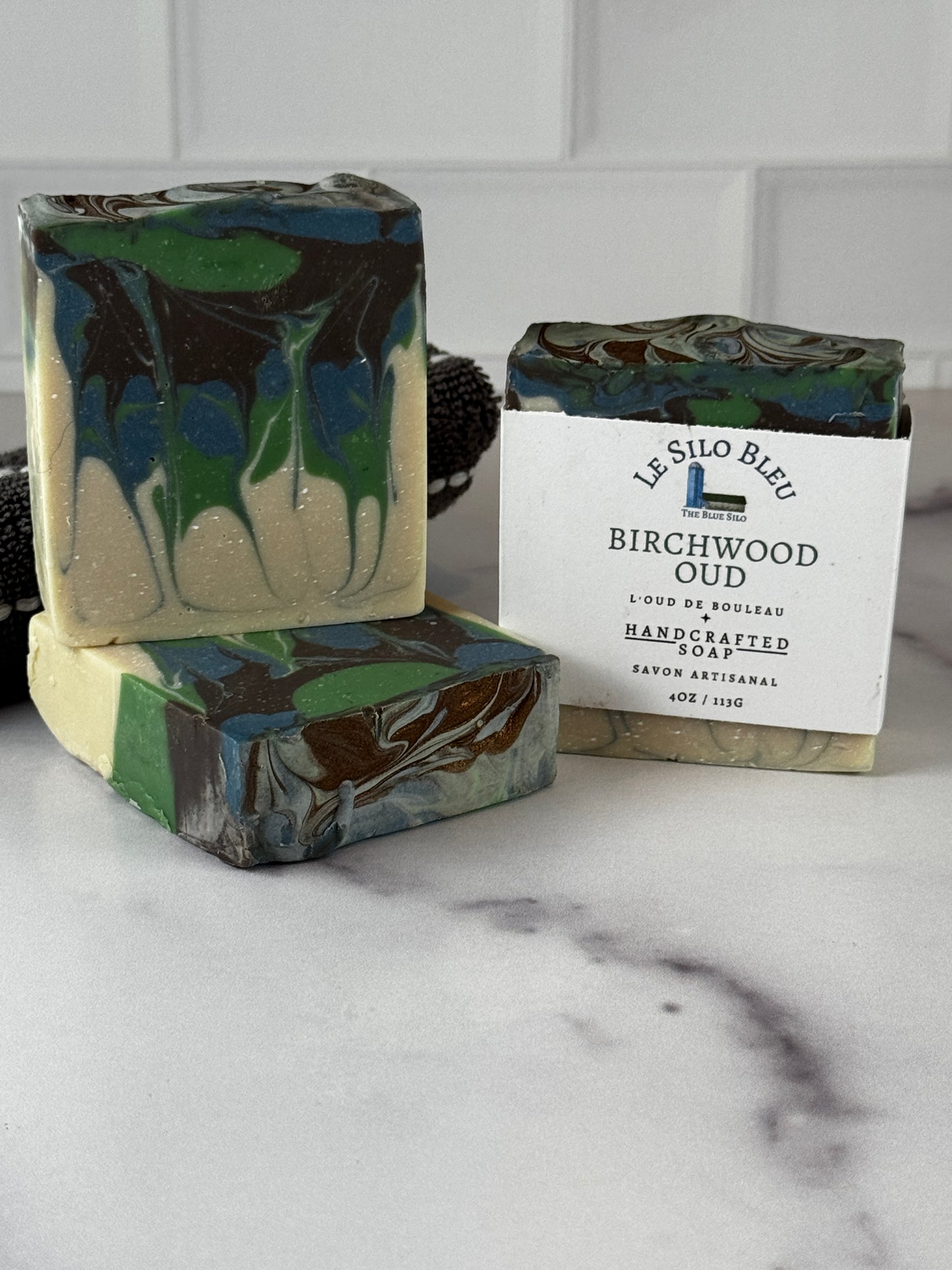 Three handcrafted marbled soaps in green, blue, cream, and dark brown tones, with one soap wrapped in a white paper label reading 'Le Silo Bleu Birchwood Oud Handcrafted Soap.' The soaps are arranged on a light marble-like surface against a white tiled background.