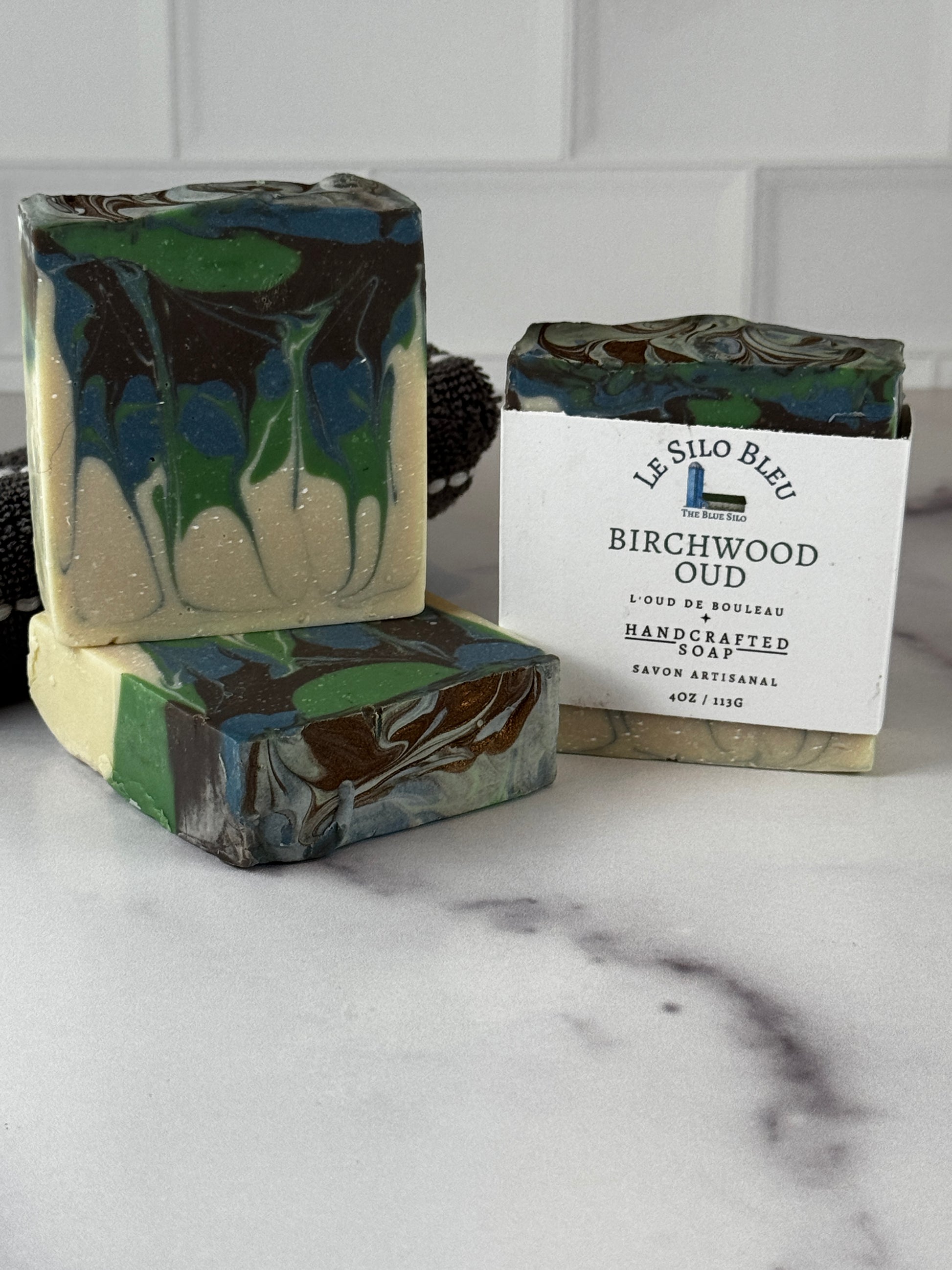 Three handcrafted marbled soaps in green, blue, cream, and dark brown tones, with one soap wrapped in a white paper label reading 'Le Silo Bleu Birchwood Oud Handcrafted Soap.' The soaps are arranged on a light marble-like surface against a white tiled background.