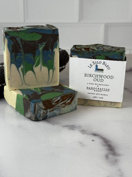 Three handcrafted marbled soaps in green, blue, cream, and dark brown tones, with one soap wrapped in a white paper label reading 'Le Silo Bleu Birchwood Oud Handcrafted Soap.' The soaps are arranged on a light marble-like surface against a white tiled background.