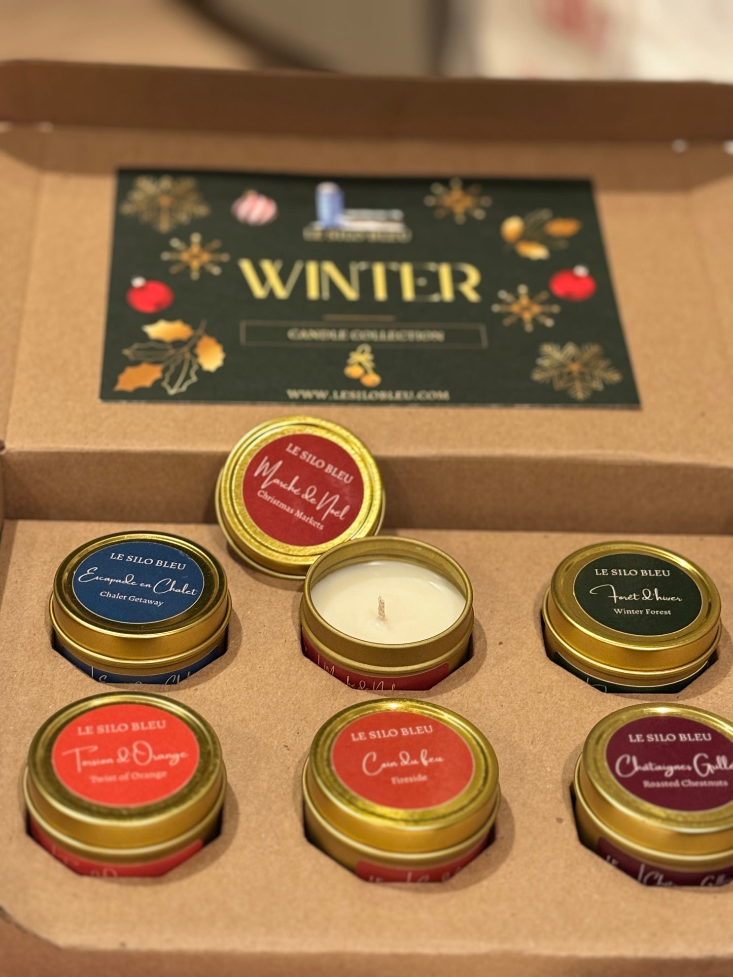 Collection of candles in gold tins with visible brand names on a 'Winter' themed box.