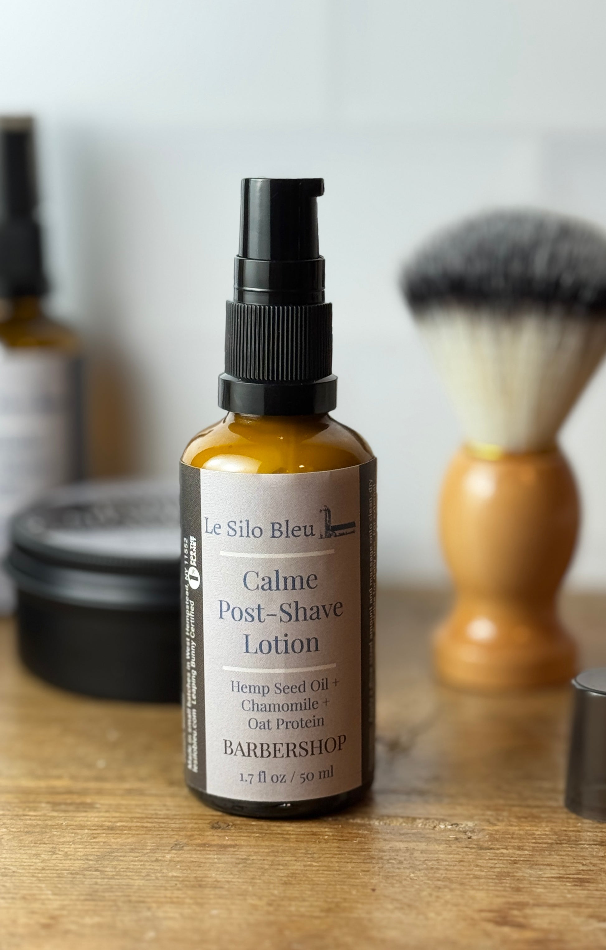 A bottle of Le Silo Bleu Calme Post-Shave Lotion stands on a wooden surface with a shaving brush and other blurred grooming items in the background.