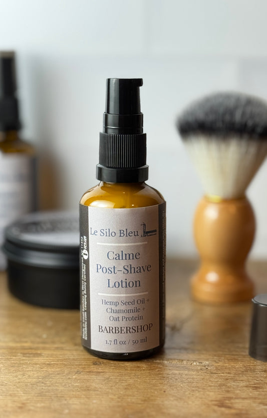 A bottle of Le Silo Bleu Calme Post-Shave Lotion stands on a wooden surface with a shaving brush and other blurred grooming items in the background.