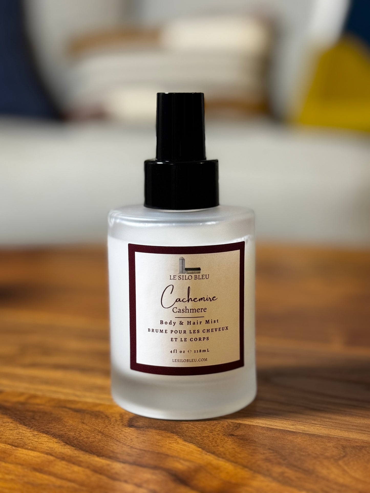 A frosted glass spray bottle of Le Silo Bleu Cashmere Body & Hair Mist with a black pump and a cream label, resting on a wooden tabletop with a blurred indoor living-room  background.
