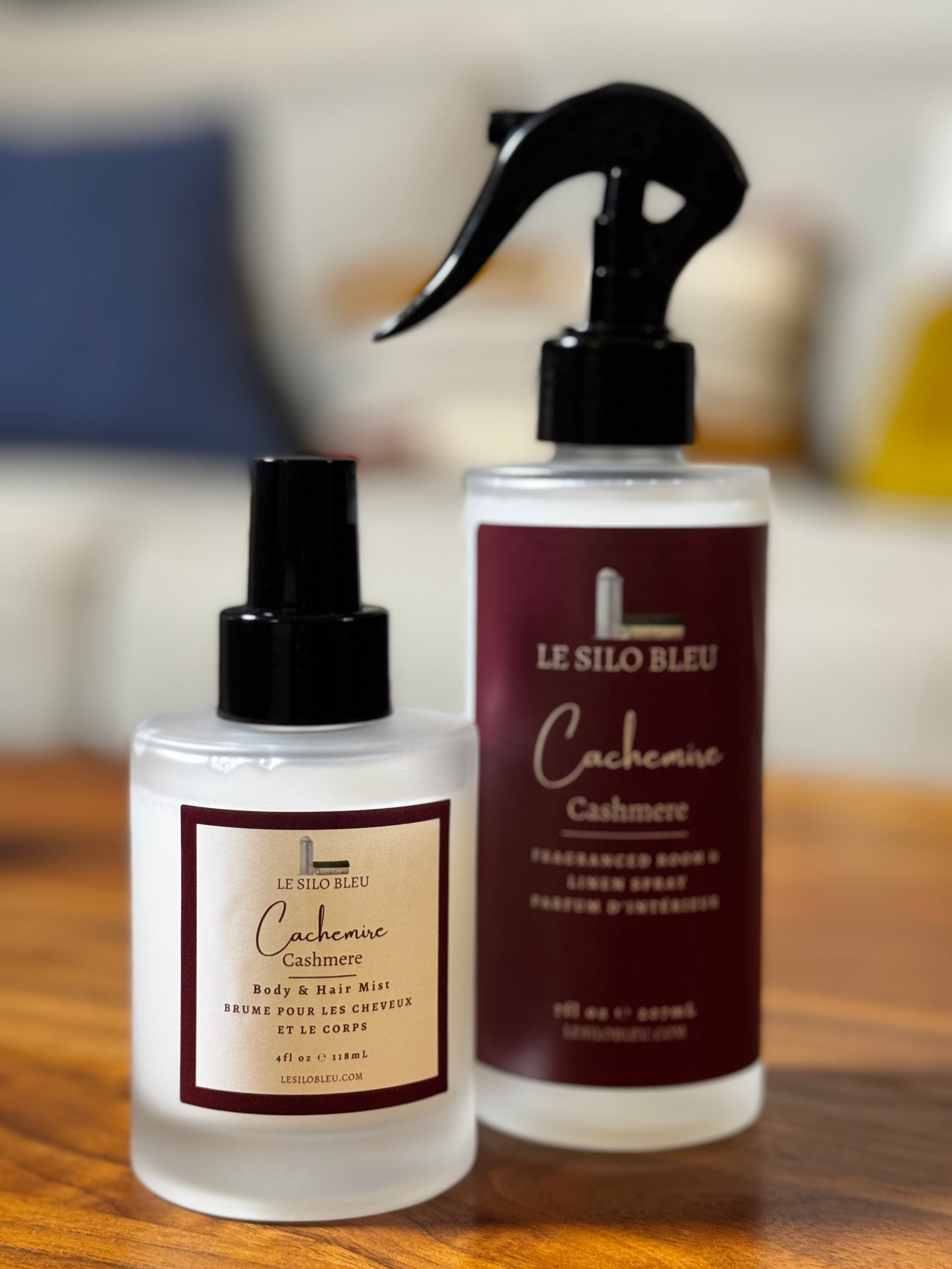 Two Le Silo Bleu Cashmere fragrance products- room spray and body mist on a wooden surface, featuring a smaller clear bottle with a black spray cap and a larger clear bottle with a black trigger sprayer and a dark burgundy label.